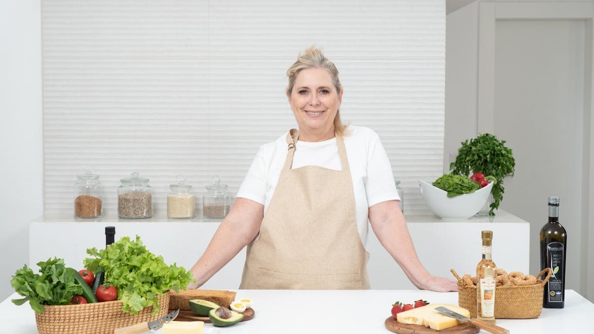 A mature woman in an apron stands at a kitchen counter with fresh produce and cheese, ready for cooking.